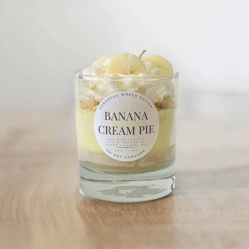 Candle in a glass jar labeled 'Banana Cream Pie' on a wooden surface.
