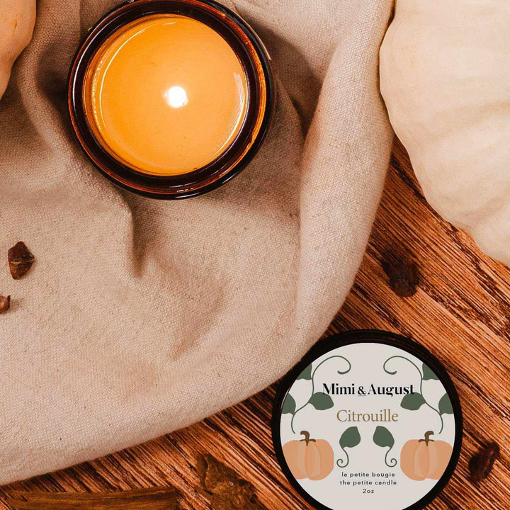 A lit amber glass jar candle sits on a beige fabric surface next to its round lid, which reads &