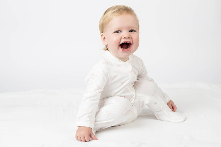 A smiling baby sitting on a white blanket wearing a white long-sleeve footed sleeper with a subtle pink pattern.