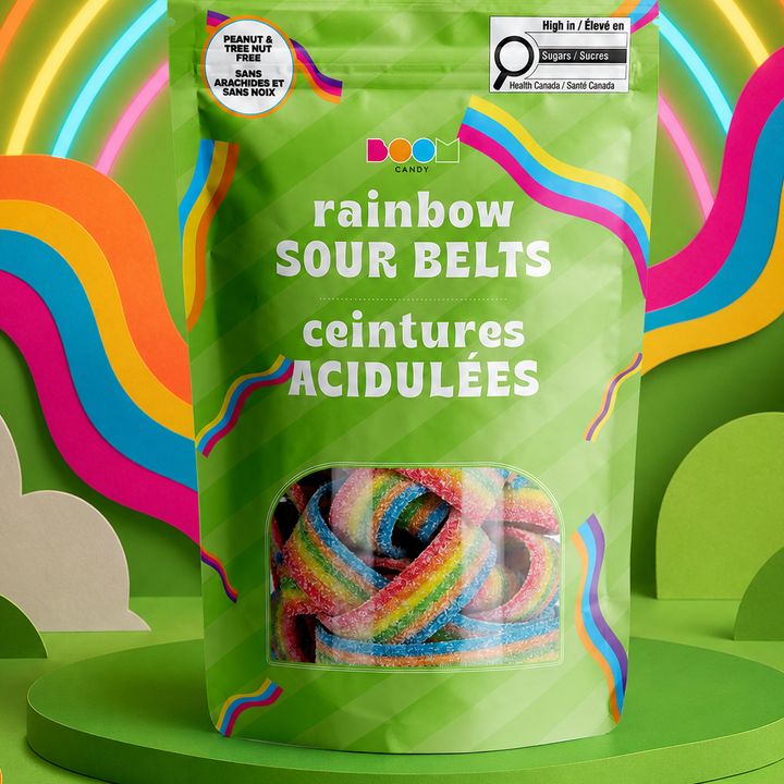 A green stand-up pouch of rainbow sour belts candy is centered on a green pedestal against a colorful illustrated background with neon rainbow arcs, wavy stripes, clouds, and stars. The front of the package reads &