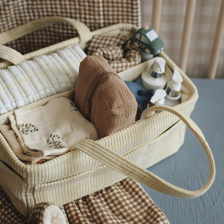 Wicker basket with baby items including a blanket, bottle, and toys on a checkered chair.