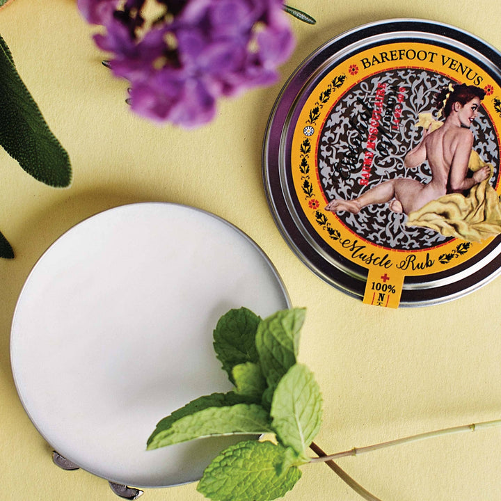 Barefoot Venus Muscle Rub container with a white disc on a yellow background with purple flowers and green leaves.