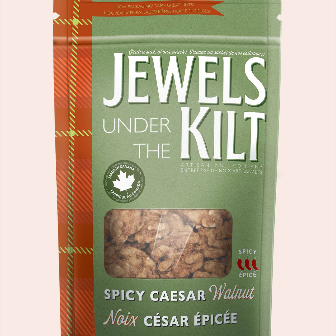 A stand-up pouch of Jewels Under the Kilt Spicy Caesar Walnut, featuring a sage green background with a red-orange tartan pattern on the left side and a clear window showing the walnut pieces inside.