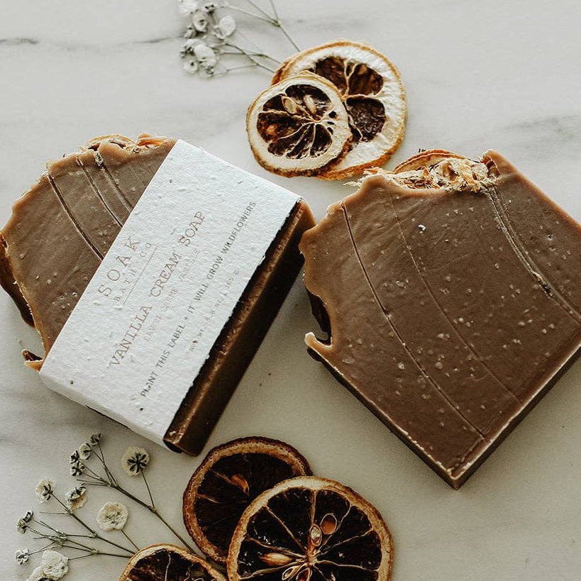 Two chocolate-colored artisanal soap bars with dried orange slices and small white flowers arranged on a light marble surface. One soap bar has a white paper band wrapped around it with the brand name &