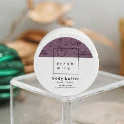 A small round tin of Fresh Wife Body Butter sits on a clear acrylic display block. The label is white with a purple graphic and black text.