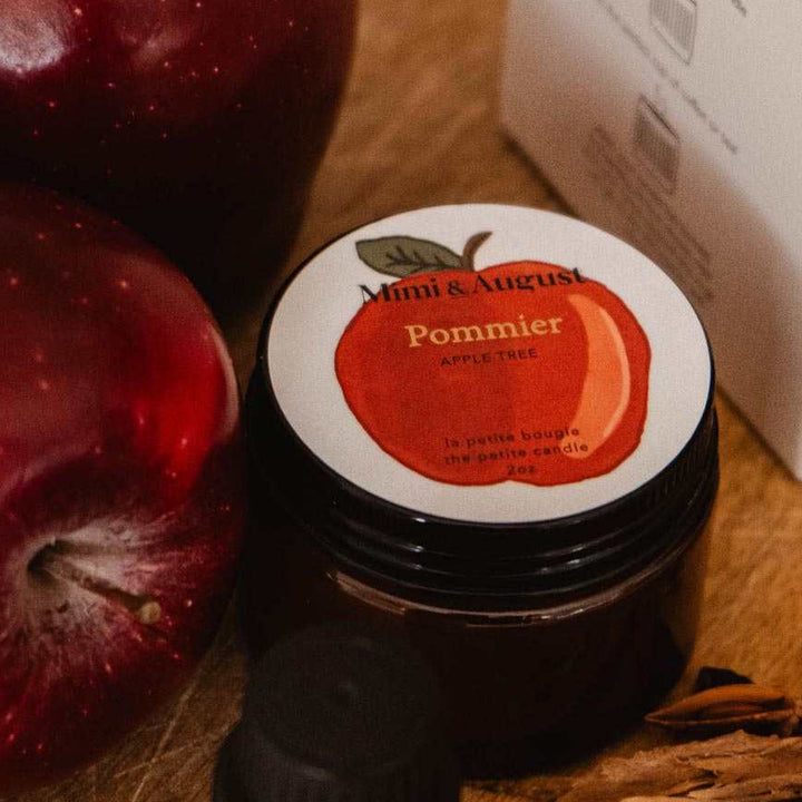 A styled product shot featuring a small bottle of essential oil, a jar of pomace, and a branded box on a wooden board, alongside two red apples and star anise. The brand &
