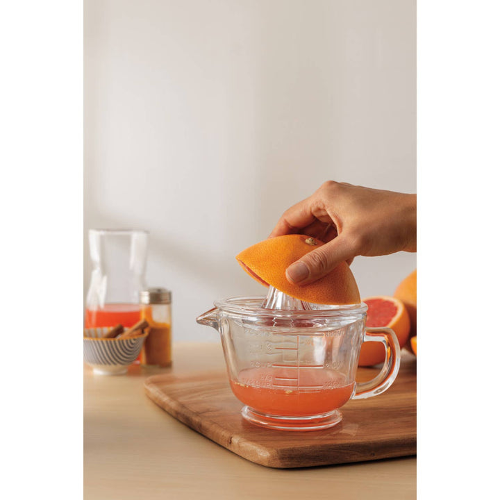 Hand using a citrus press to extract juice from an orange into a clear glass pitcher on a wooden cutting board.