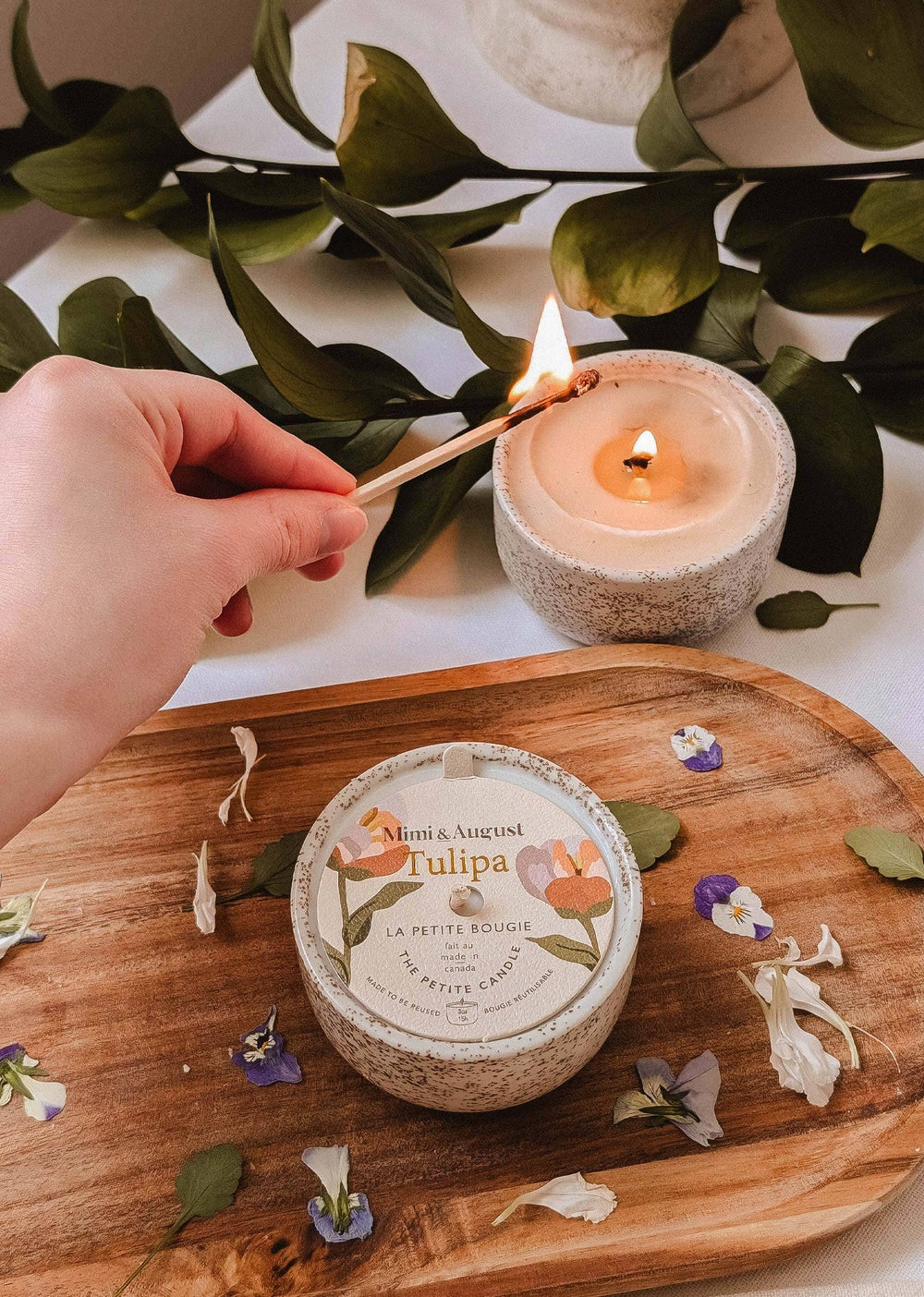 Hand lighting a candle on a wooden board with a container labeled 'Maison et Jardin' and greenery in the background.