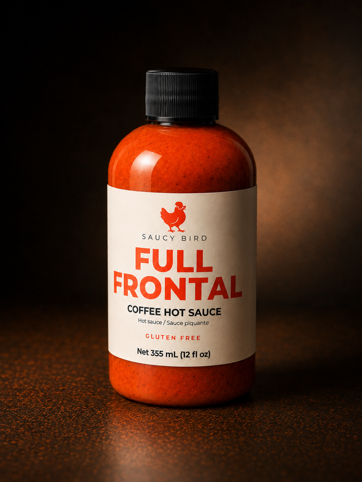 A bottle of Saucy Bird Full Frontal Coffee Hot Sauce, filled with a vibrant orange-red sauce, featuring a white label with red and black text, and a black cap, set against a dark, moody background.