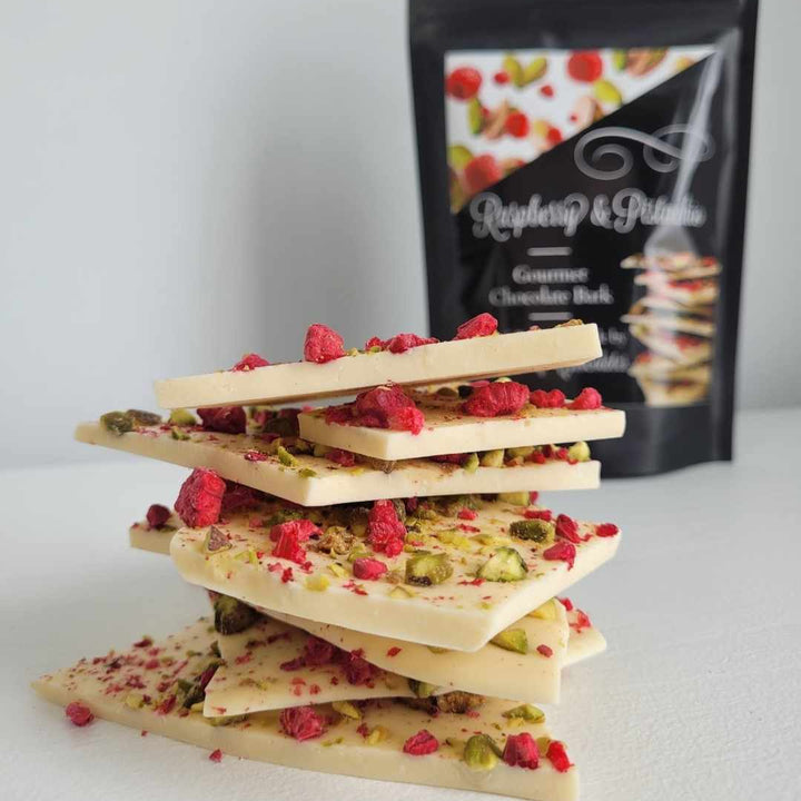 A stack of white chocolate bars topped with chopped pistachios and freeze-dried raspberries, with a packet of product in the background.
