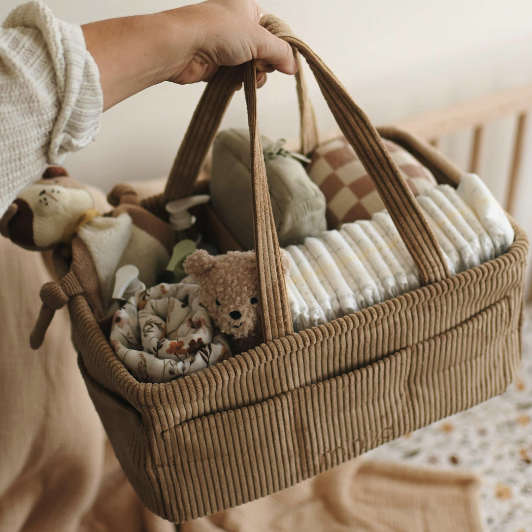 Brown woven basket filled with baby items including stuffed animals, held by a person in a soft, warm-toned room.