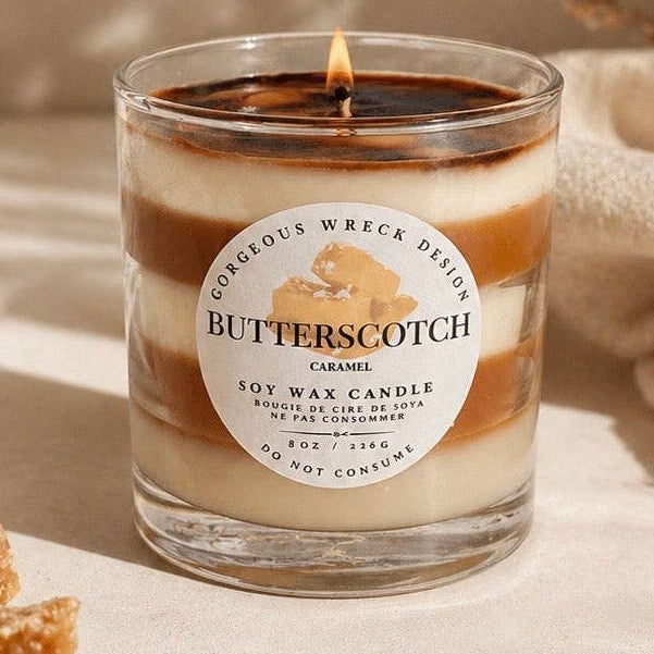 Butterscotch candle with a label on a beige surface with caramel pieces and dried leaves.