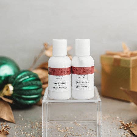 Two small white hand lotion bottles with white caps and red-and-white labels, displayed on a clear acrylic stand. The scene is styled with green ornaments, dried leaves, and a wrapped gift box in the background.