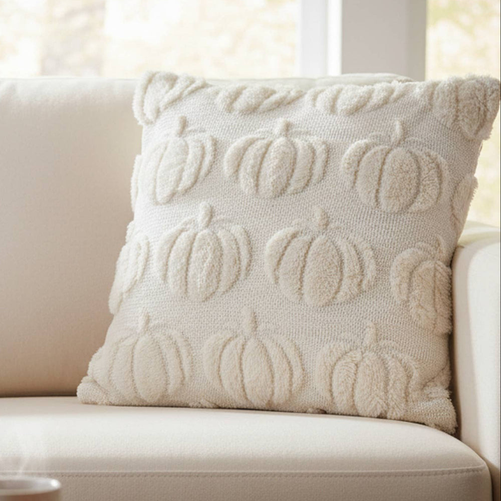 An off-white textured throw pillow with a raised pumpkin pattern styled on a cream sofa in a cozy living room setting, with a wooden coffee table, a mug, and a bowl of fruit in the foreground.