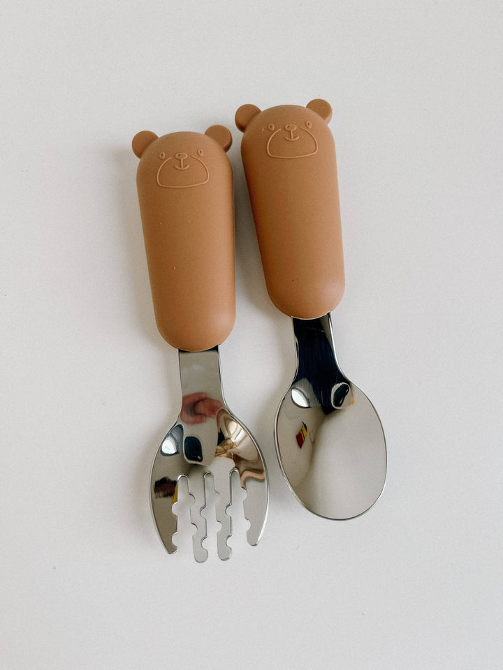 A baby utensil set featuring a brown bear-shaped spork and a matching brown bear-shaped spoon with stainless steel bowls, displayed on a clean white background.