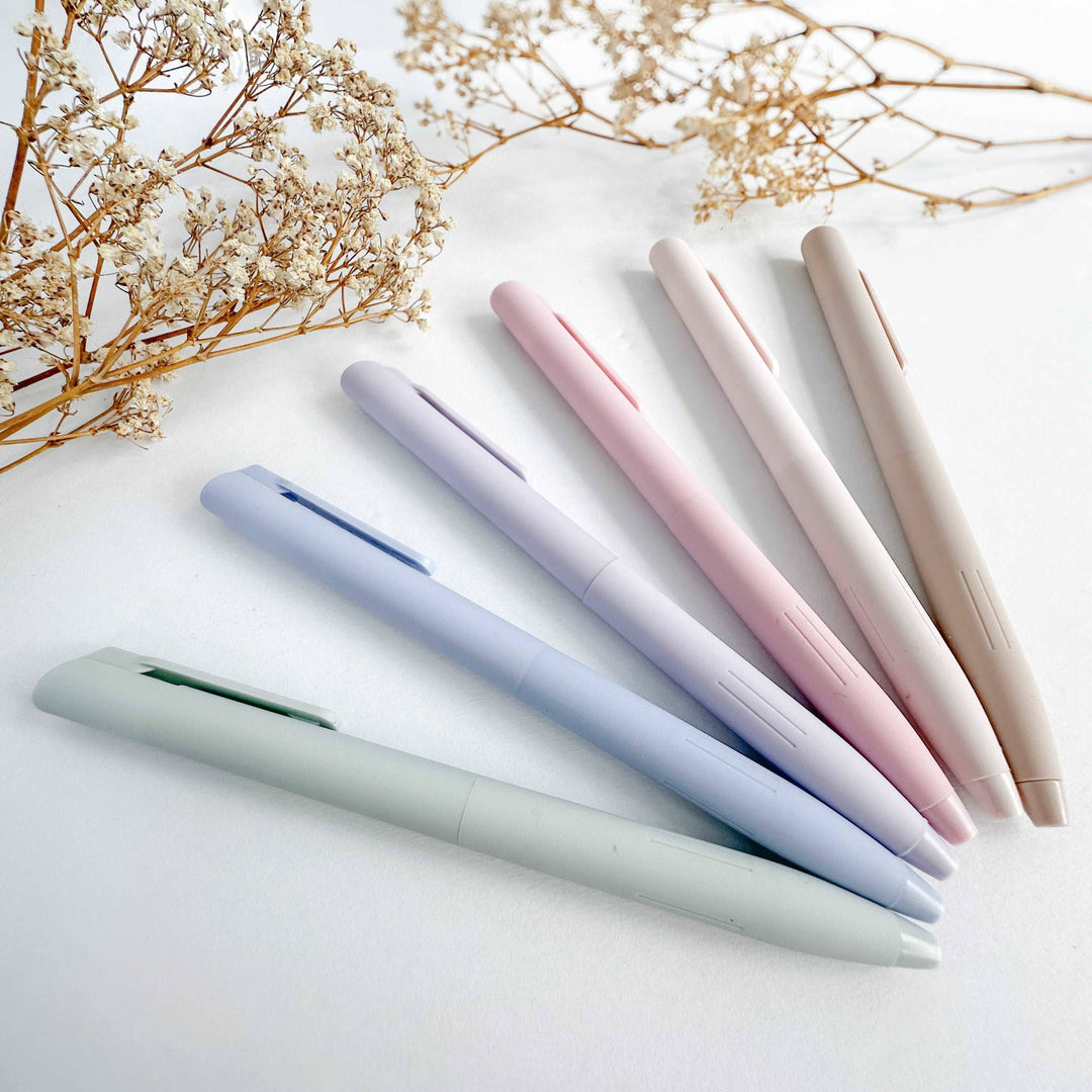 Six pastel-colored ballpoint pens in shades of sage green, lavender, light blue, pink, cream, and beige, arranged diagonally on a white surface with dried baby&