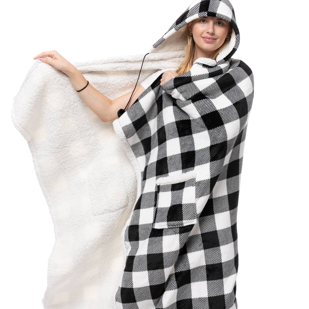 A woman modeling a hooded blanket throw with a black and white buffalo plaid exterior and a soft white sherpa lining, holding the open flap of the blanket with one hand.