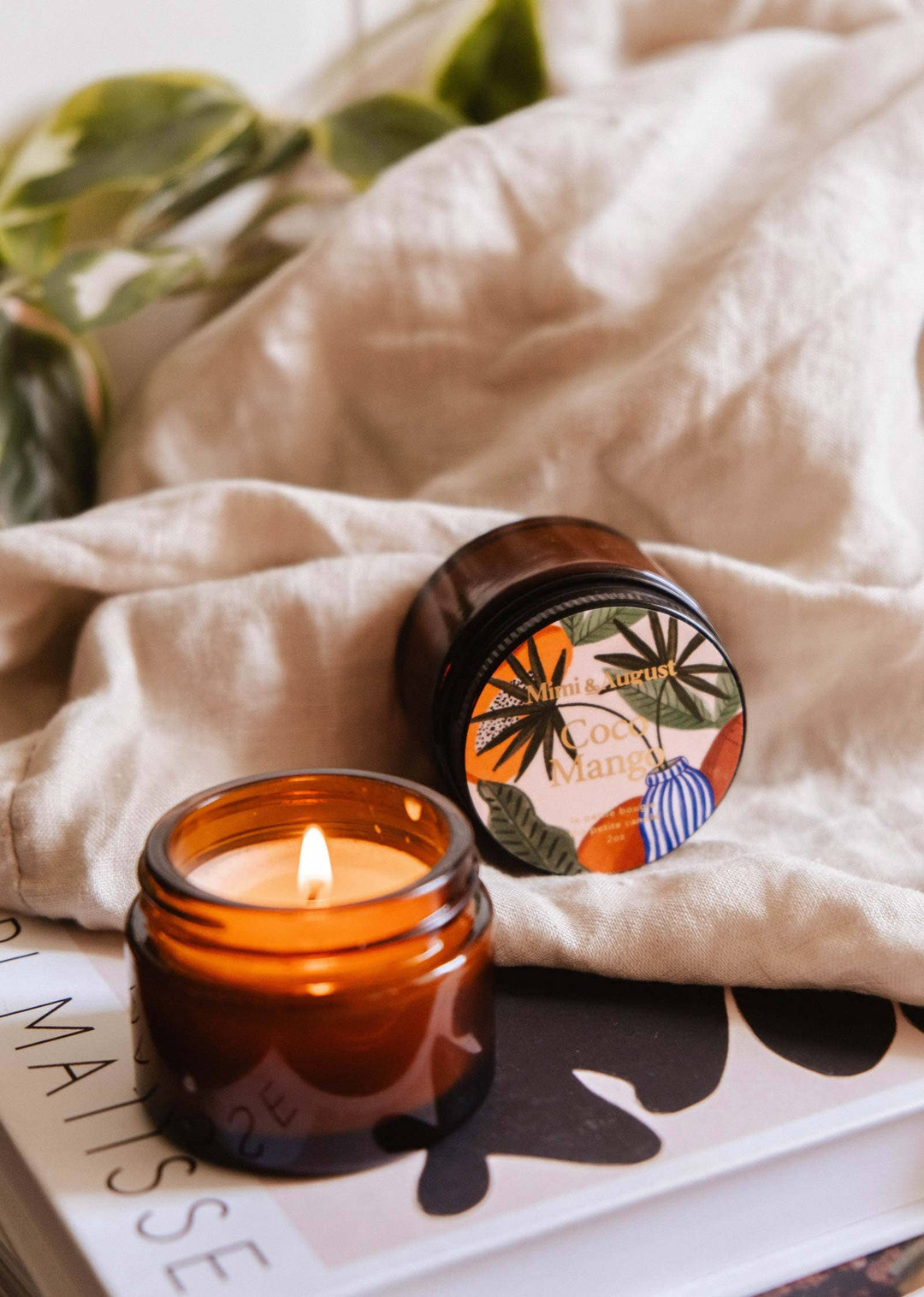 Candle in an amber jar with a decorative lid on a magazine, surrounded by beige fabric and green leaves.
