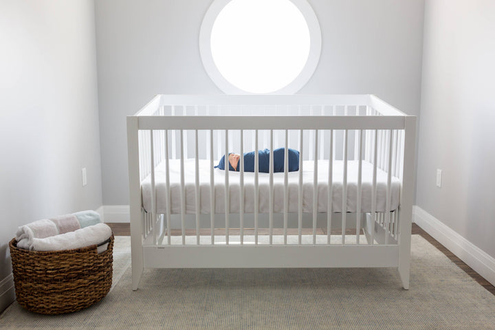 White crib with a baby inside in a room with a round window and a basket on the floor.