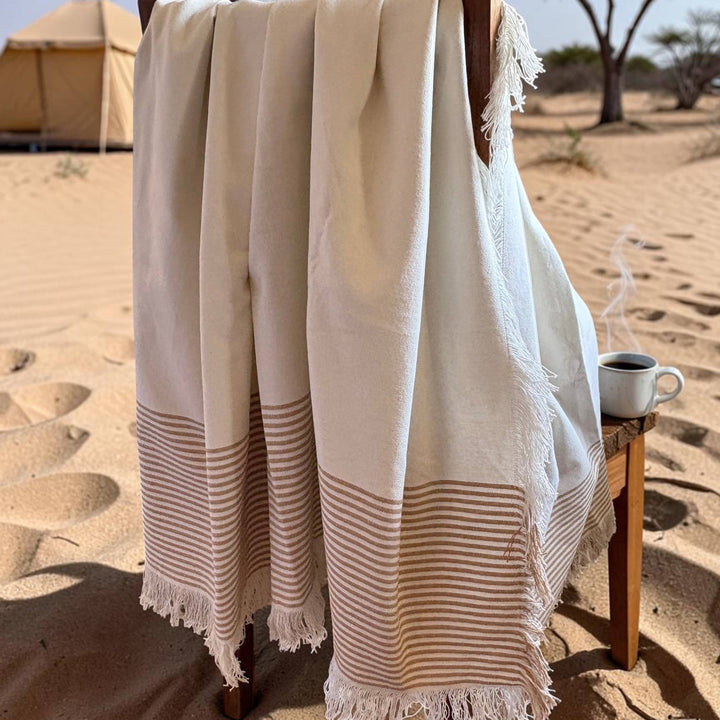 A cream and beige striped cotton towel with fringed edges draped over a wooden chair in a sandy desert setting, with a small table holding a steaming mug nearby.