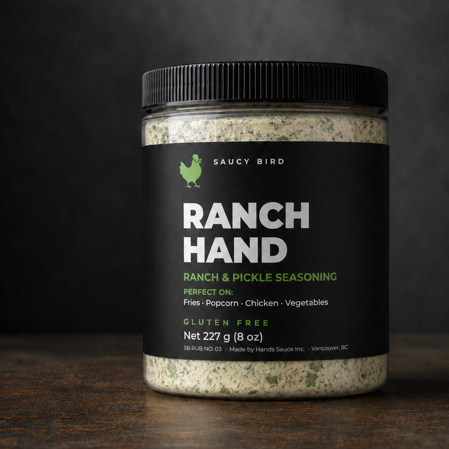 A jar of Saucy Bird Ranch Hand Ranch &amp; Pickle Seasoning sits on a dark wooden surface against a dark, smoky background. The product is in a clear plastic jar showing the beige and green seasoning blend inside, with a black lid and a black label featuring white and green text.
