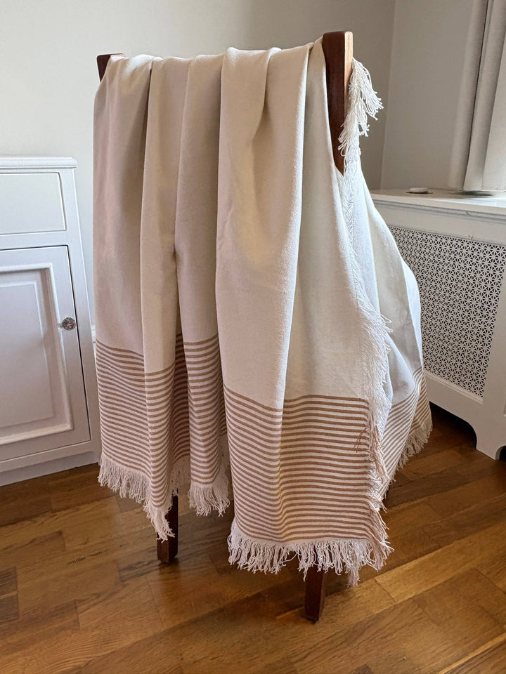 A cream-colored Turkish cotton throw blanket with fringed edges and a striped pattern near the bottom, draped over a wooden chair in a home interior setting.