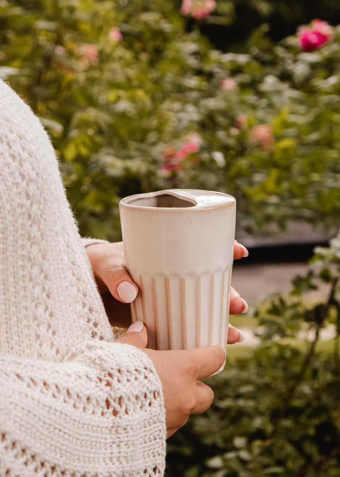 A person in a cream knit sweater holds a beige ceramic travel mug with vertical fluting and a small cutout lid, set against a soft-focus garden background with green foliage and pink flowers.