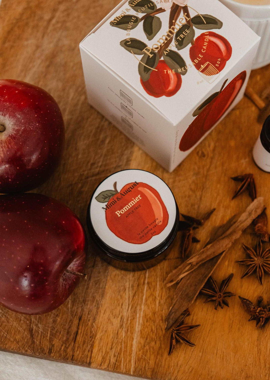 A small round jar of Romier apple cider vinegar with a red apple illustration label, shown next to its white box decorated with red apples, on a wooden cutting board with whole apples, cinnamon sticks, and star anise.