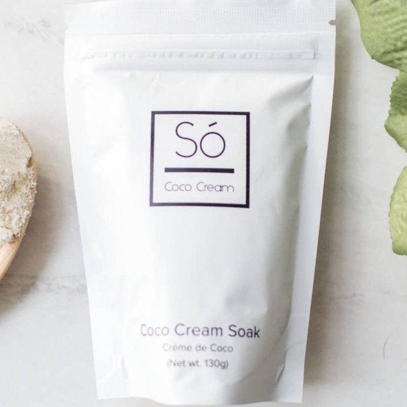 Só Coco Cream packaging on a light surface with a wooden spoon and plant leaves.