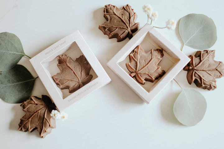 Two square white gift boxes containing leaf-shaped soap bars, arranged on a white surface with decorative eucalyptus leaves and small white flowers. The soap bars are brown and shaped like maple leaves.