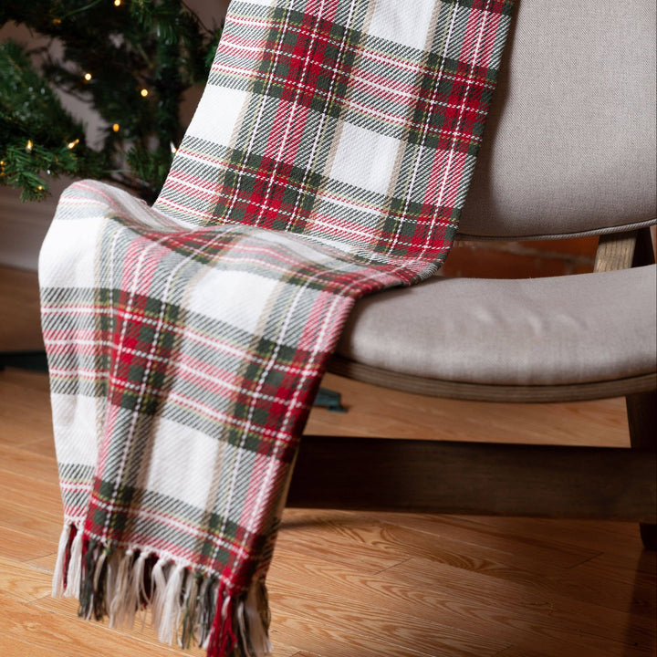 A plaid throw blanket draped over a light gray upholstered chair in a living room setting, with a Christmas tree and warm wood floor visible in the background.