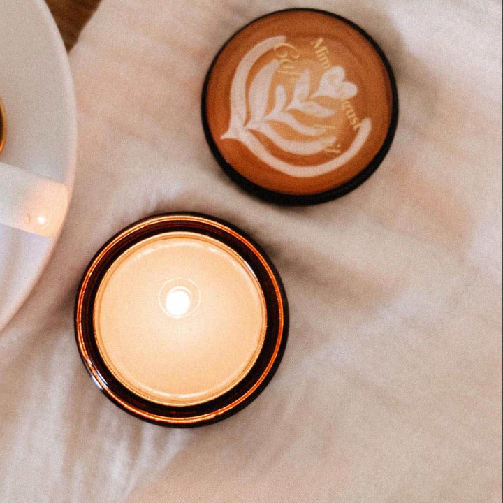A cozy tabletop scene with a cup of coffee on a saucer, a plate of croissants, a folded cream cloth, and two small amber glass candle jars, one lit and one showing latte art.