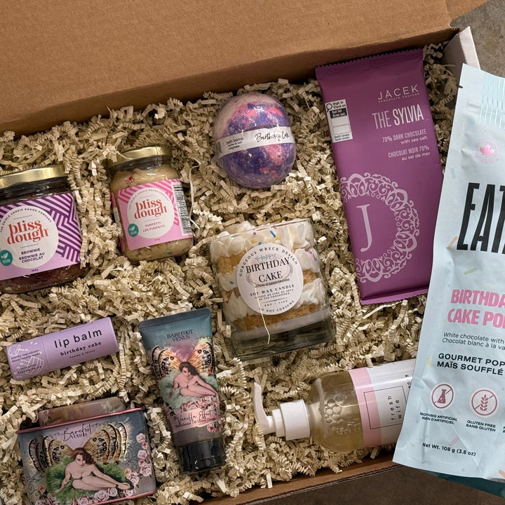 A birthday gift box with cookie dough, birthday cake flavoured lip balm, lotion & bath salts by Barefoot Venus, a Jacek chocolate bar, birthday bath bomb, birthday cake candle, and birthday cake pop popcorn by Eatable. 