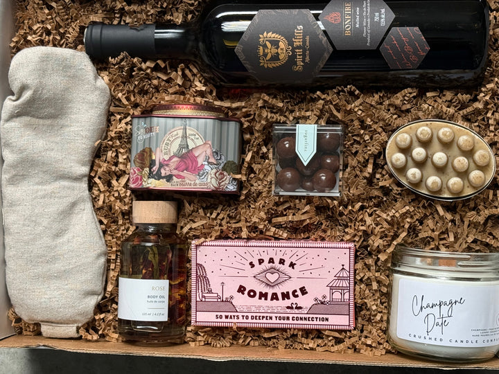 Gift box with various items including a bottle of wine, candles, and a card on a wooden surface.