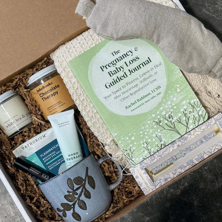 A curated gift box set shown from above, containing a sage green guided journal, a gray ceramic mug with a leaf pattern, a candle, a jar of chocolate therapy, a hand cream tube, a candlestick, and a gray cloth, all arranged in brown crinkle paper inside a white box.