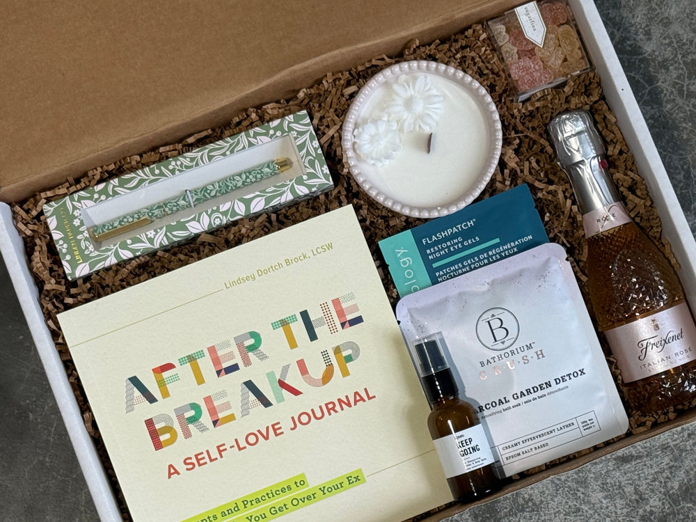 A curated gift box set containing a cream-colored self-love journal titled 'After the Breakup', a white candle, a boxed item with green botanical patterns, a small bottle, a white bath and body product pouch, and a bottle of Prosecco, all arranged in brown packing paper inside a white box.