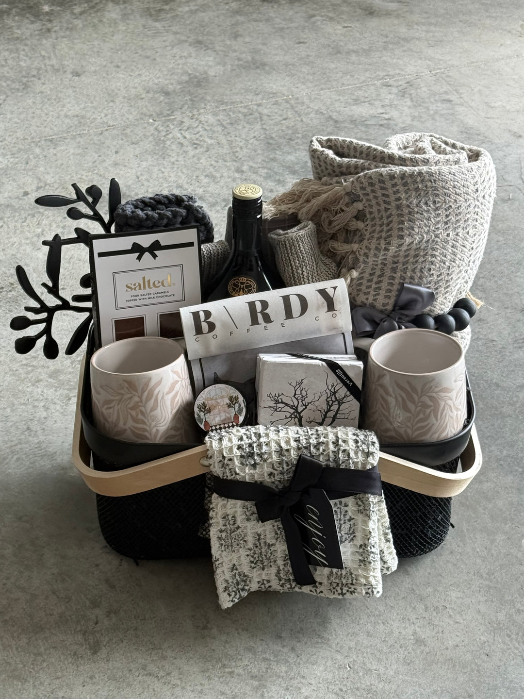 A curated gift basket or hamper arranged in a light wood tray on a gray concrete floor. The set includes a folded knit throw blanket, ceramic mugs, a patterned fabric pouch tied with a black ribbon, printed cards, and a dark woven textile.