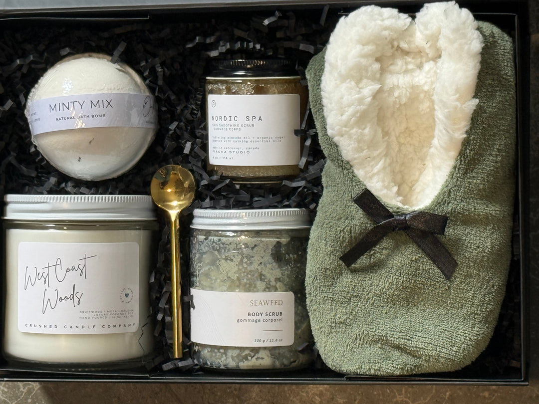 A curated spa gift set in a black box with shredded black paper filler, featuring a round bath bomb labeled &