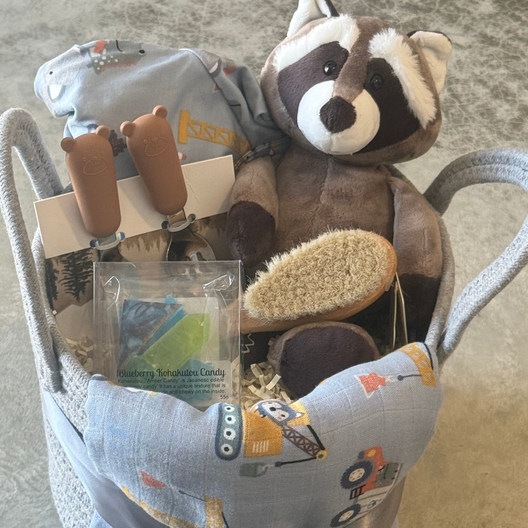 A baby gift basket filled with toys and baby items, including a plush raccoon, a soft toy, a brush, a small packaged item, and a light blue fabric basket with construction-themed print and a gray ribbon bow.