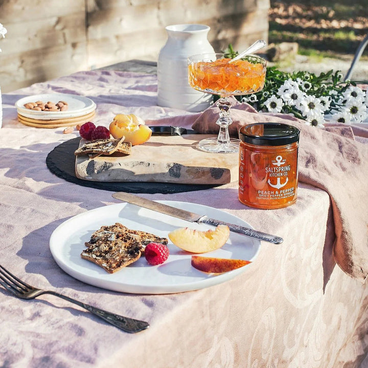 An outdoor dining setup with a plate of food, including what appears to be a piece of pizza, fruits, and cheese, accompanied by a jar of Peach & Pepper Spicy Pepper Spread.