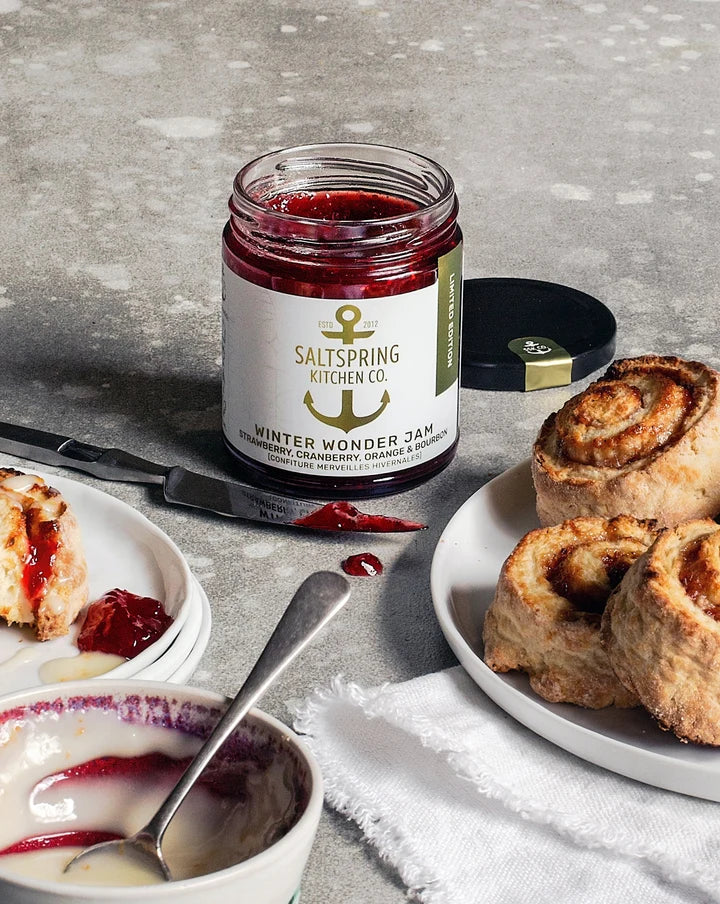 A jar of Saltispring Kitchen Co. Winter Wonder Jam sits on a gray stone surface, surrounded by plates of cinnamon rolls and jam spread, with the lid placed beside the jar.