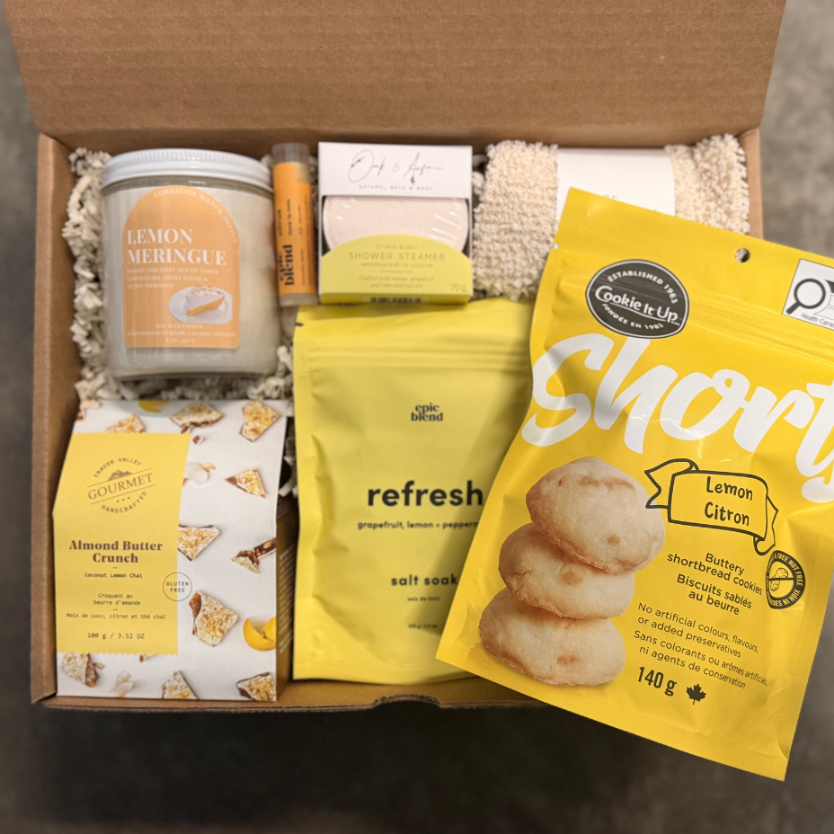 Open box containing various items including a candle, cookies, and snacks on a neutral background