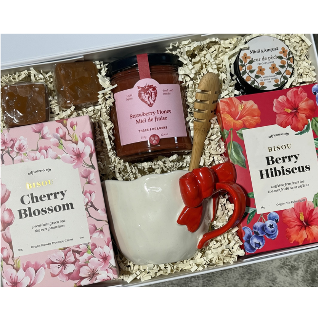 A curated gift box containing a jar of Berry Jam, a pink Cherry Blossom soap, a box of Berry Hibiscus soap, a white ceramic mug with a red ribbon, and a wooden cinnamon stick, all arranged in crinkle paper.