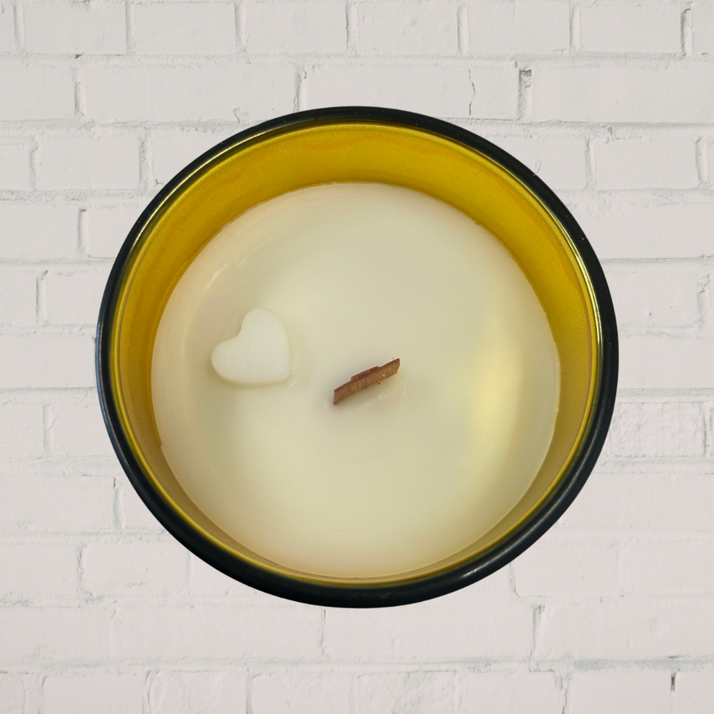 Candle with a heart-shaped wick in a black and gold container against a white brick wall.