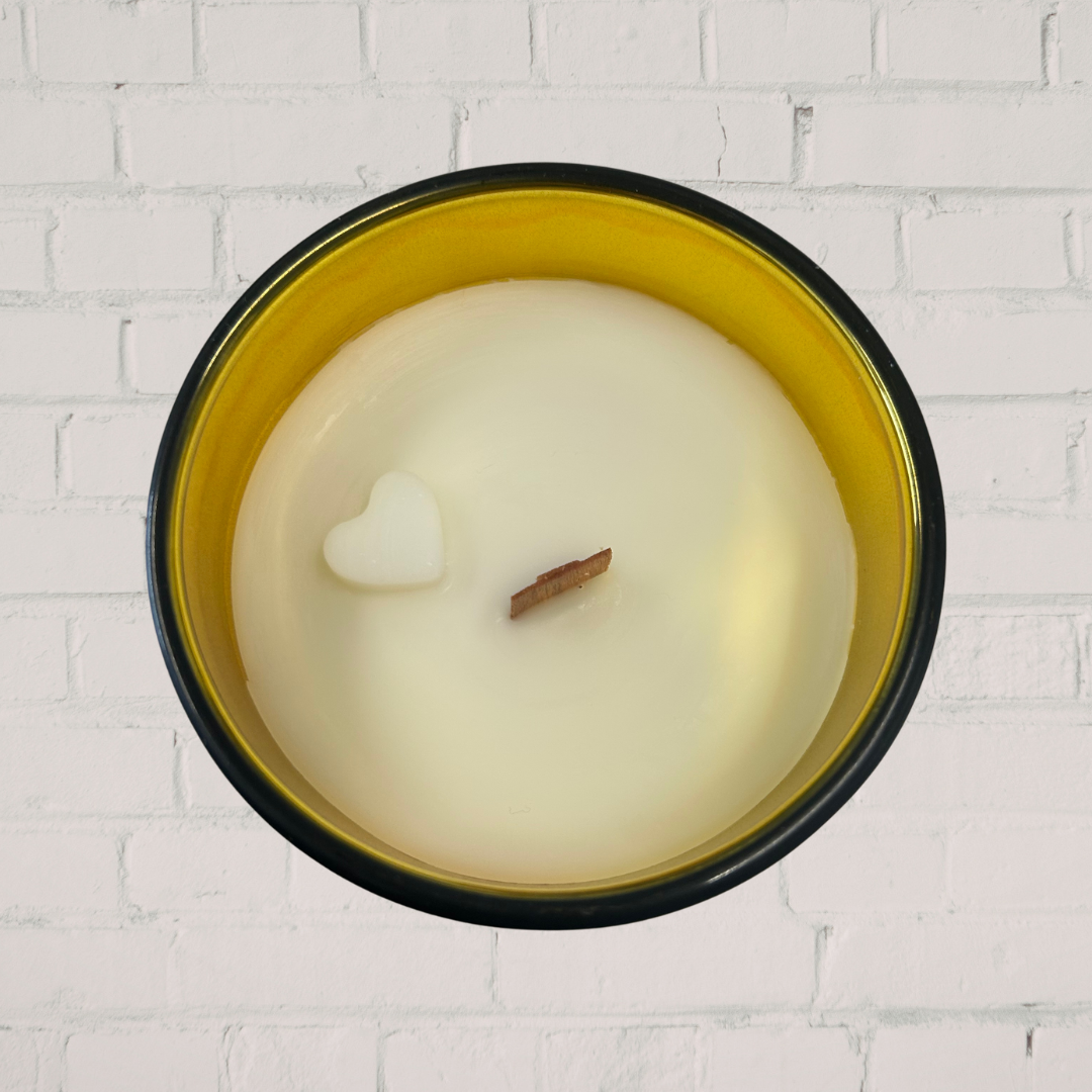 Candle with a heart-shaped wick in a black and gold container against a white brick wall.