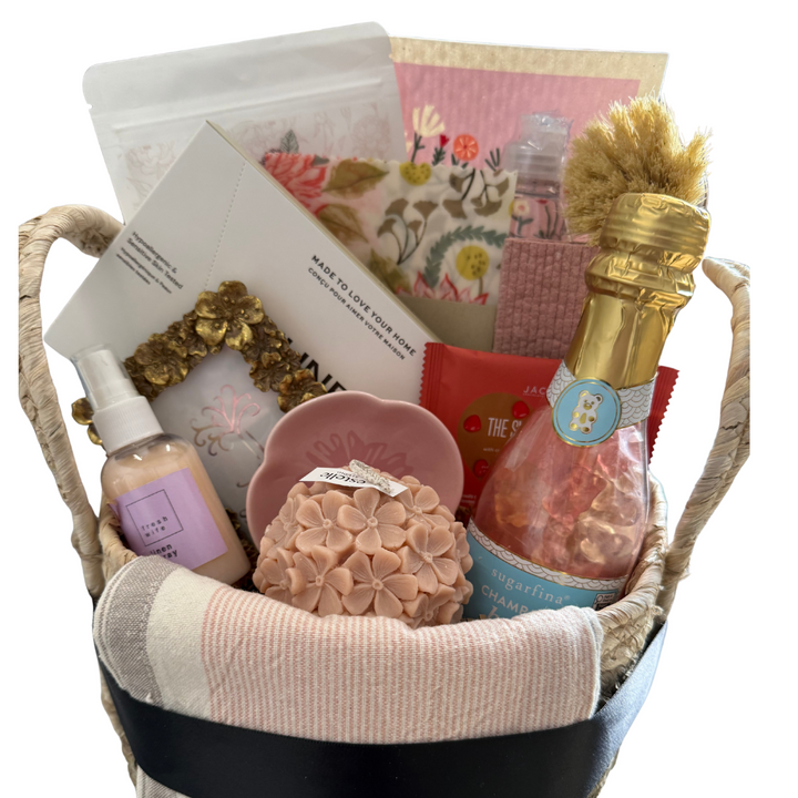 Housewarming gift basket in blush, floral, gold and white including candles, swedish dish cloths, dryer sheets, linen spray, champagne gummy bears and dish towels.