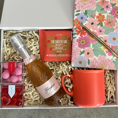 Gift box with a bottle of champagne, mug, chocolate, candies, and stationery items on a textured surface
