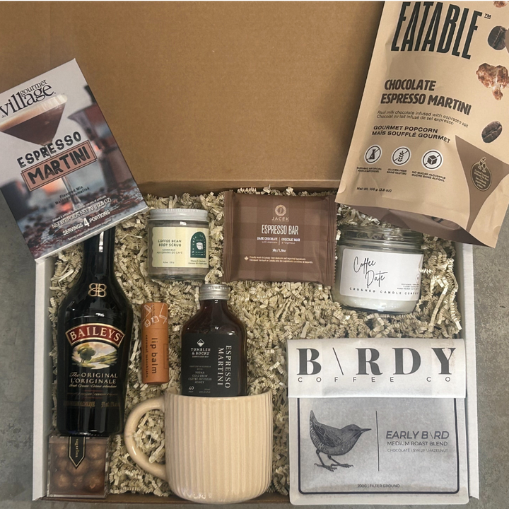 Gourmet gift box with coffee-themed items including a bottle, mug, and packaging on a gray surface.