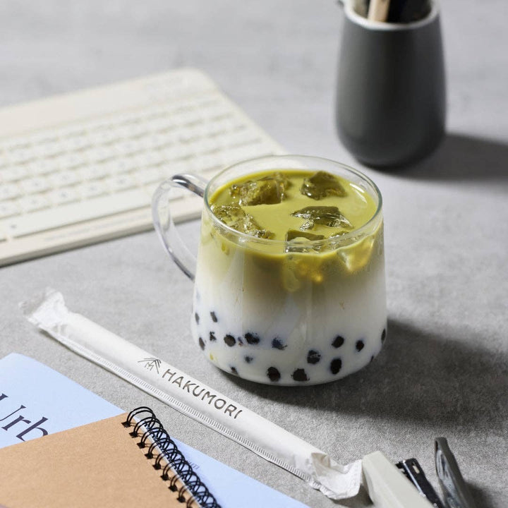 Green iced drink with boba in a clear mug on a desk with a Makumori straw.