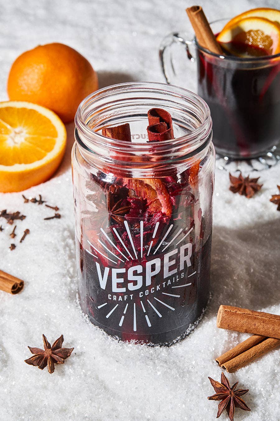 Vesper craft cocktails jar with mulled wine, cinnamon sticks, and oranges on a textured surface.